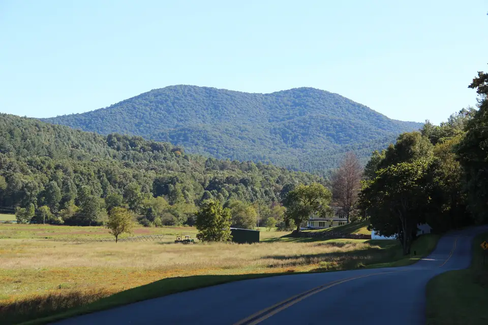 Forested double-summit mountain under a clear blue sky above a sunlit hay meadow, with a winding two-lane road in the foreground and scattered trees and a farmhouse.
