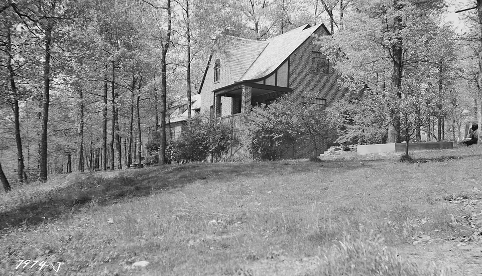 Black-and-white photograph of a brick Tudor-style house with steep gables and a stone porch among slender trees on a grassy slope, with a handwritten negative number at lower left and a dark film border.