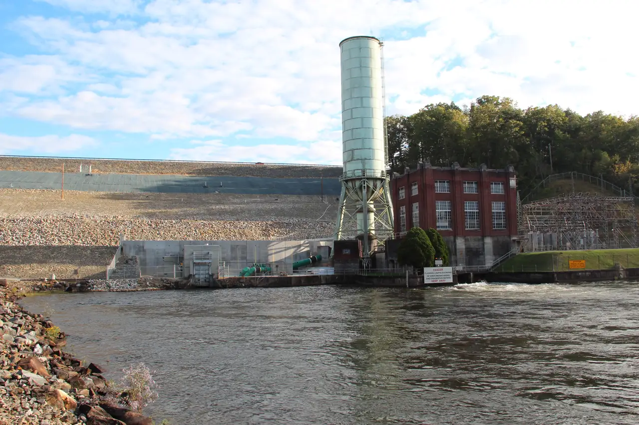 Rock-faced earth embankment of the dam with a tall cylindrical surge tank on a steel frame, a red-brick powerhouse with tall windows, switchyard structures at right, and the river flowing out in the foreground past rock riprap.