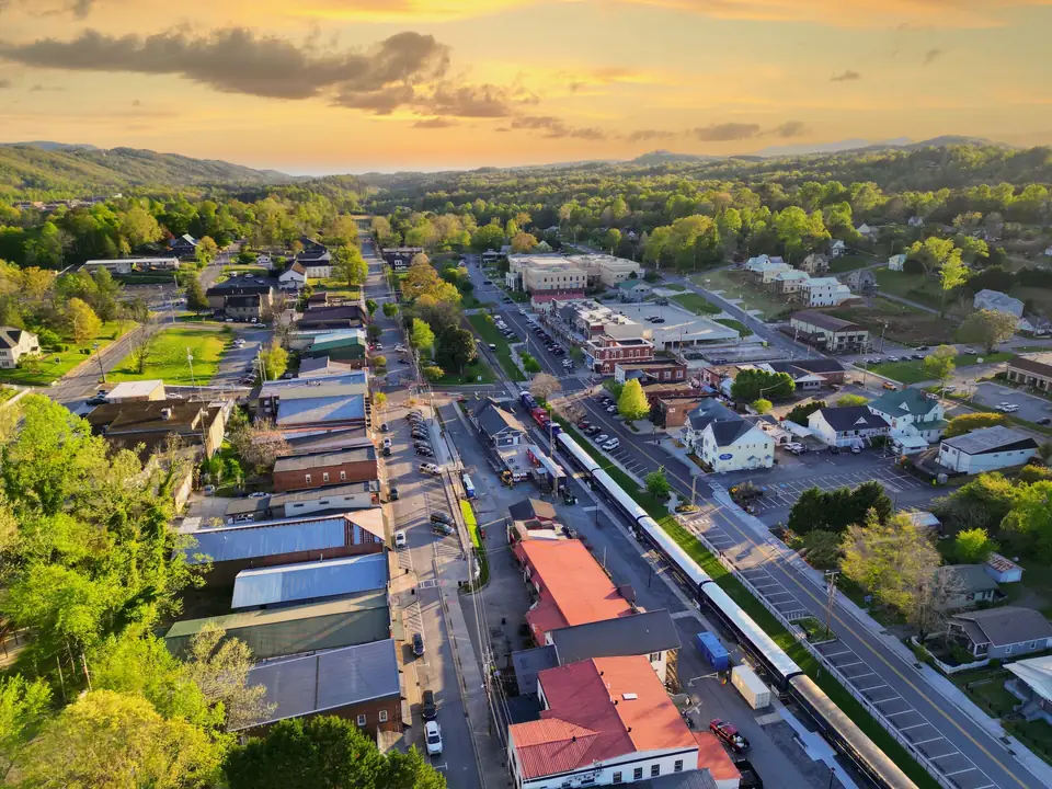 Drone view over the downtown blocks with red, blue and grey metal roofs, the scenic railway train beside a parking area, tree-lined streets and green hills rising to a sunset sky.