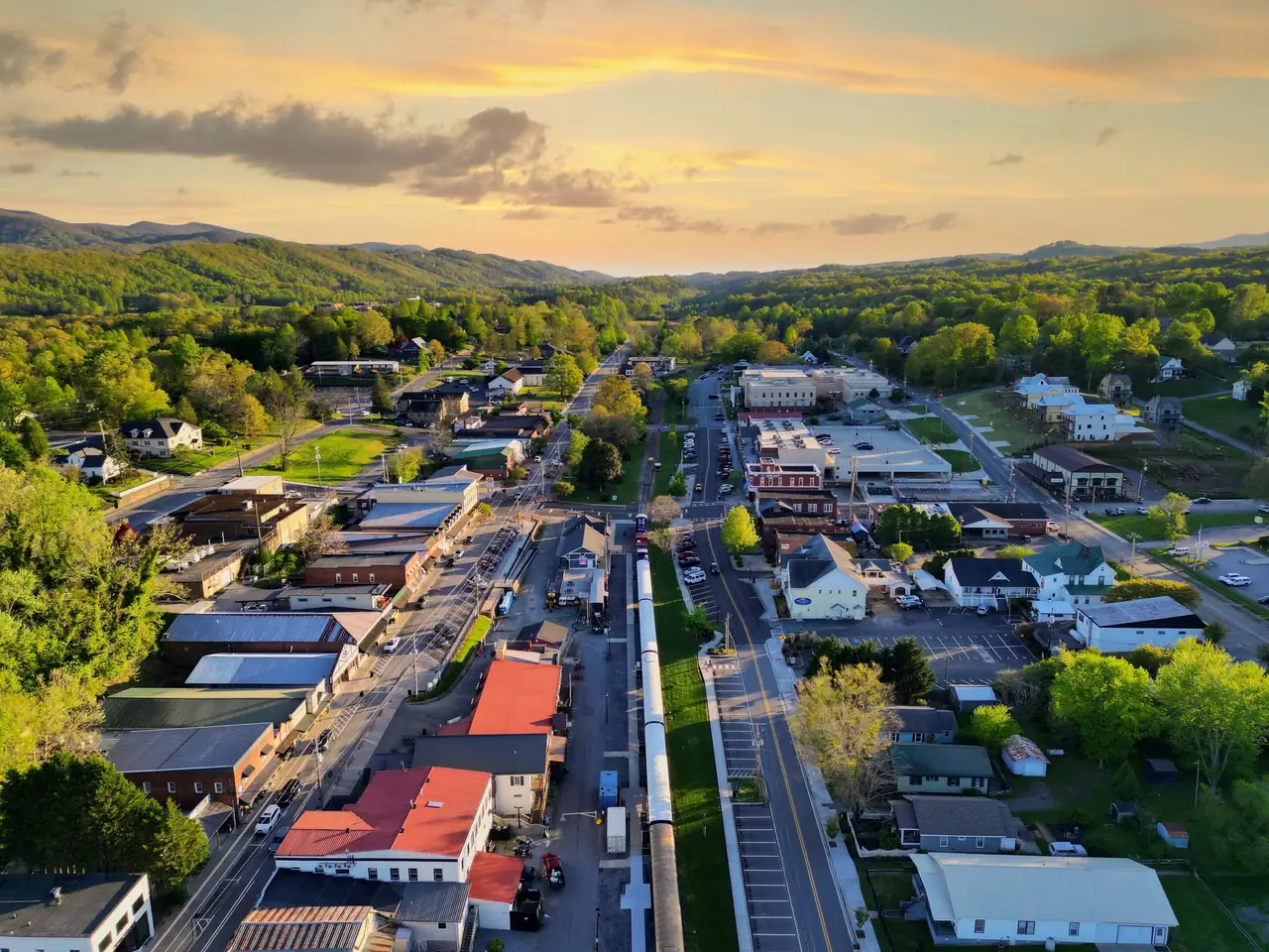 Drone view down the downtown corridor with the passenger train on the tracks, red and blue roofs, houses among green trees and long blue mountain ridges under a soft orange evening sky.