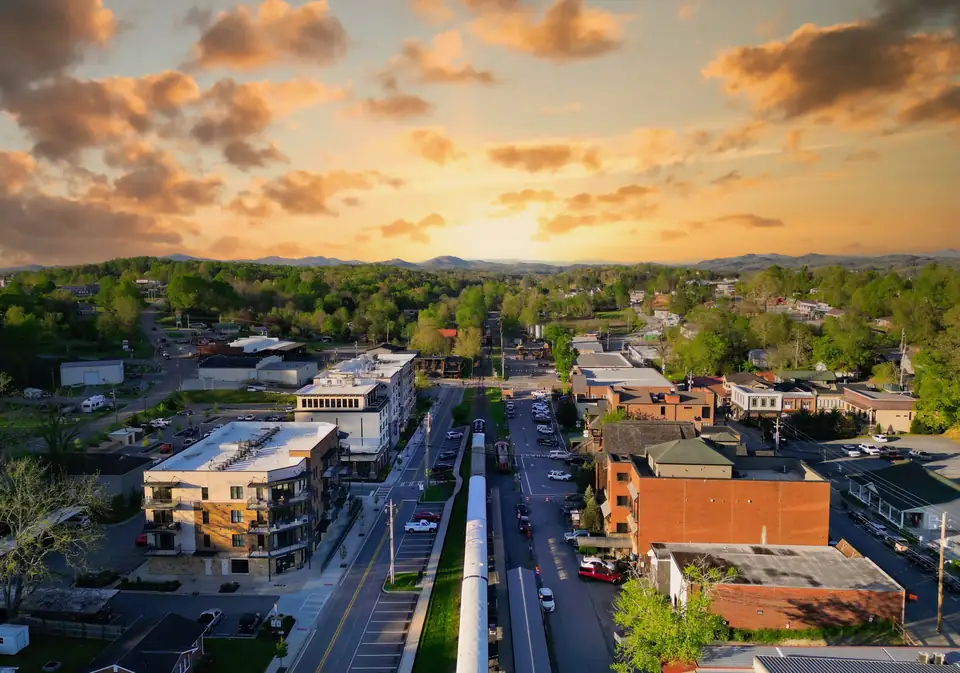 Drone view looking straight down the railroad tracks with a long white passenger train between two downtown streets, new multi-story buildings and brick blocks on either side, green trees and distant mountains under orange clouds.