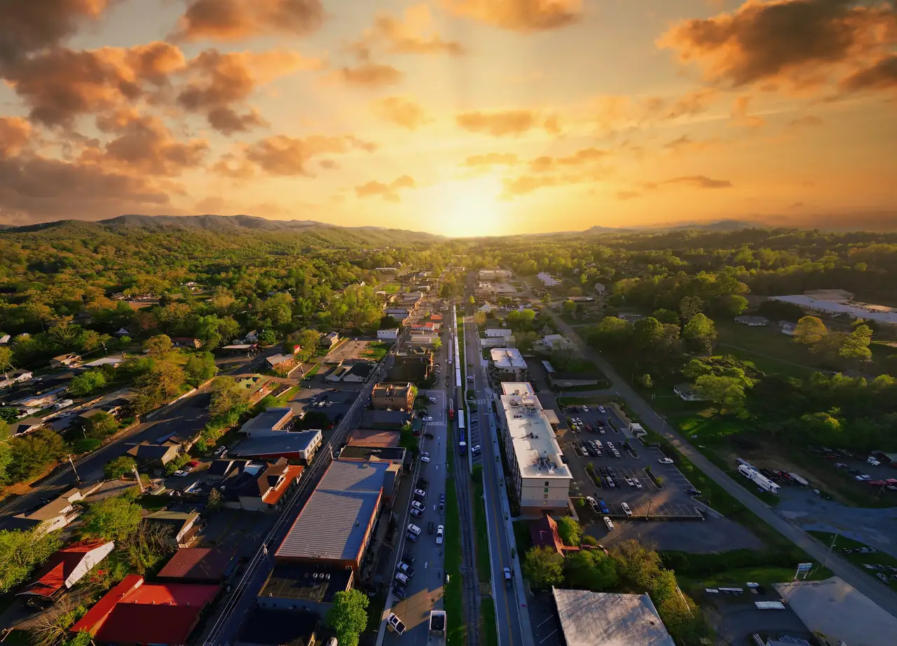 High drone view of the town grid and rail line leading toward a glowing sun setting over a forested valley, with golden rays, orange clouds and a green ridge on the horizon.
