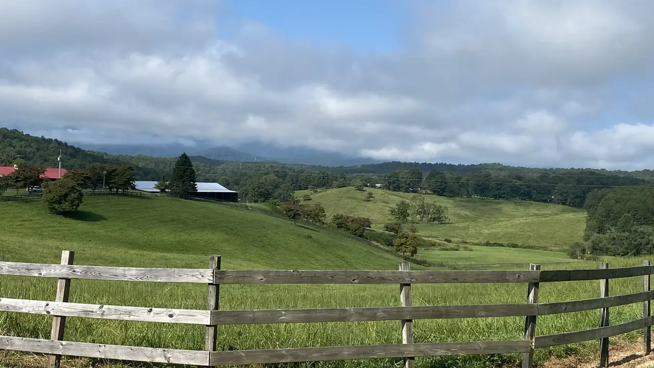 Green rolling pasture behind a weathered split-rail fence, red-roofed barns and farm buildings on a hill, wooded ridges and low cloud on the mountains beyond.
