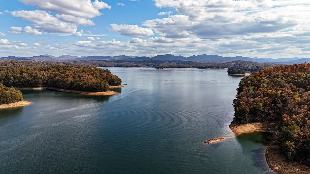 Elevated view down a long arm of the lake between wooded peninsulas in orange and brown fall color, sandy red-clay shoreline at low water, calm blue water with a faint boat wake, and layered blue mountains on the horizon under scattered white clouds.