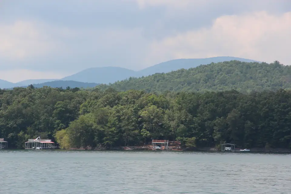 Calm lake water in front of a densely forested shoreline with several covered boat docks, a green wooded ridge rising behind, and two hazy blue mountain summits under a grey-pink sky.