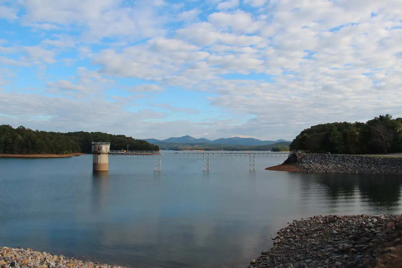 Calm blue lake behind riprap rock banks, a concrete intake tower connected by a long footbridge on piers, wooded points on both sides and a line of blue mountains across the water under a sky of scattered white clouds.