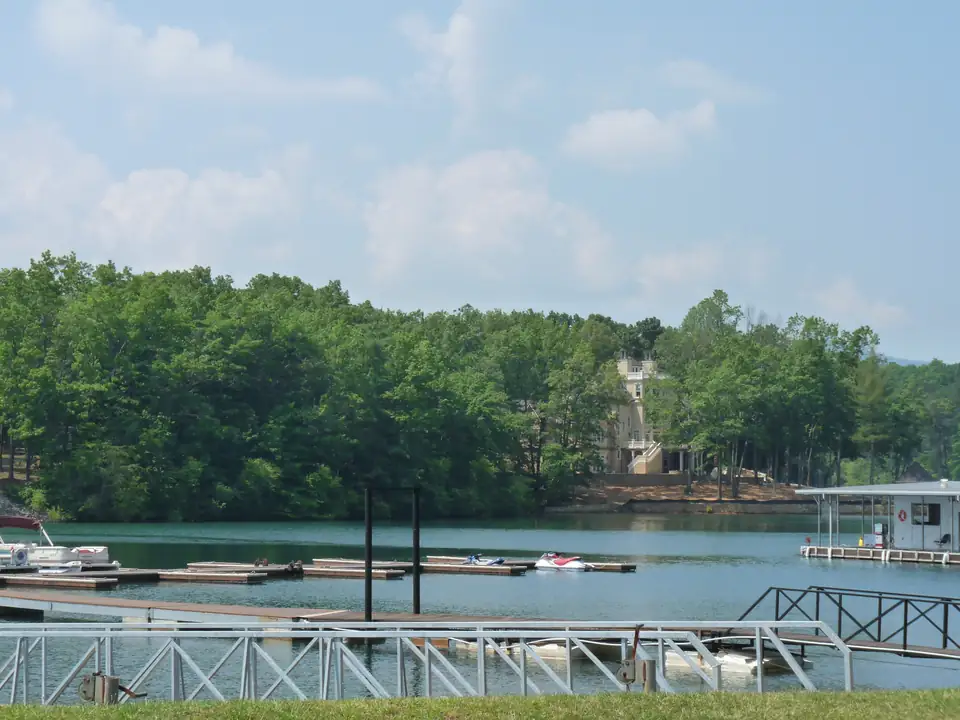 Marina boat slips and floating docks with pontoon and personal watercraft on green water, a white metal gangway railing in the foreground and a wooded hillside with one tall house behind under a hazy blue sky.