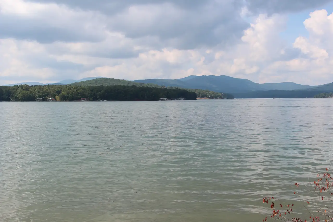 Wide expanse of green-blue open water with gentle ripples, a low forested far shore with a few docks, and rounded blue-green mountains behind under puffy white and grey clouds; a few red leaves at lower right.