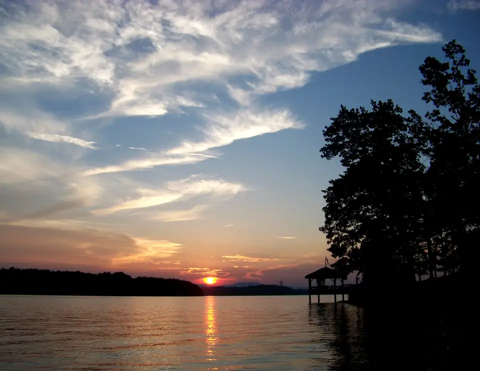 Orange sun setting on a low far shoreline with its reflection on dark water, streaky white clouds in a blue sky, and a silhouetted tree and covered boat dock on the right.