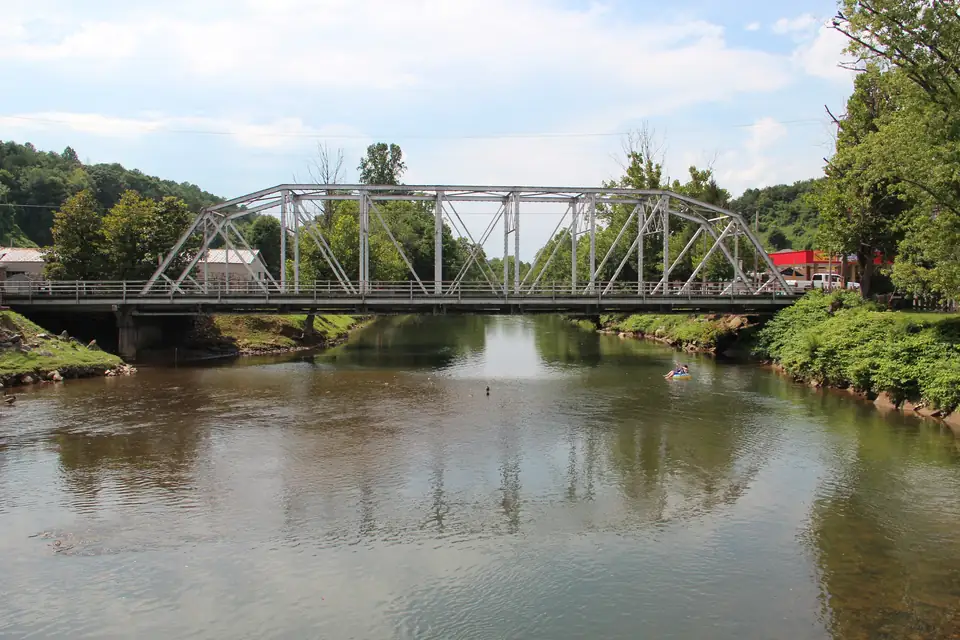 Silver steel truss bridge crossing a calm green river reflecting the bridge, a small kayaker on the water, wooded hills, low buildings and a red storefront on the right bank under blue sky with white clouds.
