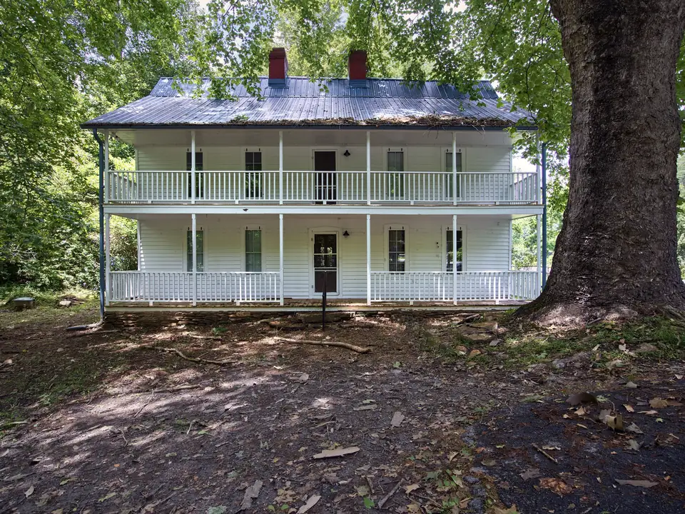 Two-story white frame house with full-width double porches, a blue metal roof and two red brick chimneys, set among tall shade trees with a huge trunk at right and a bare dirt yard in front.