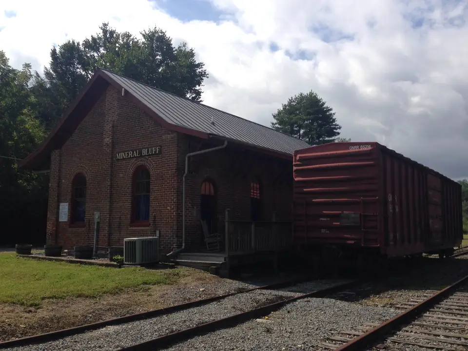 Small red-brick depot with arched windows, a green metal roof and a 'Mineral Bluff' name board, a rusty boxcar beside it on the track, grass in front and tall trees behind under a cloudy sky.