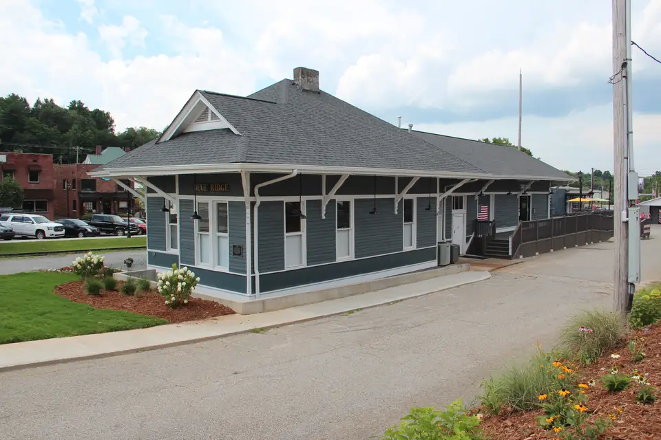 Restored blue-grey wooden depot with a hipped grey roof, wide bracketed eaves and white trim, flower beds and a lawn in front, and a parking lot and trees behind under a hazy sky.