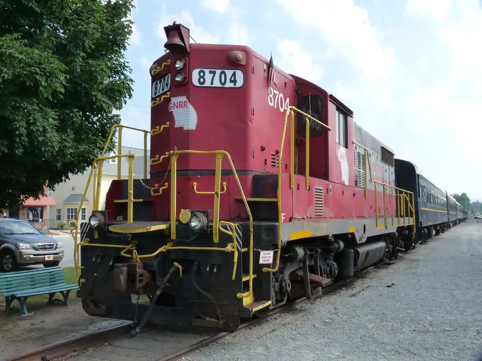 Red diesel locomotive numbered 8704 with yellow handrails at the head of a line of blue passenger coaches on gravel track, a green bench and trees at left under a pale sky.