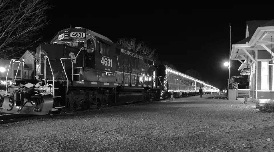 Black-and-white night photograph of a diesel locomotive lettered Blue Ridge Scenic Railway leading lighted passenger coaches beside the depot platform, with a street lamp and bare trees.