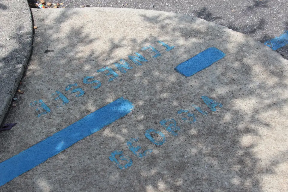 Close-up of a concrete sidewalk and curb with a painted blue line and the stenciled words GEORGIA and TENNESSEE on either side, in dappled shade.