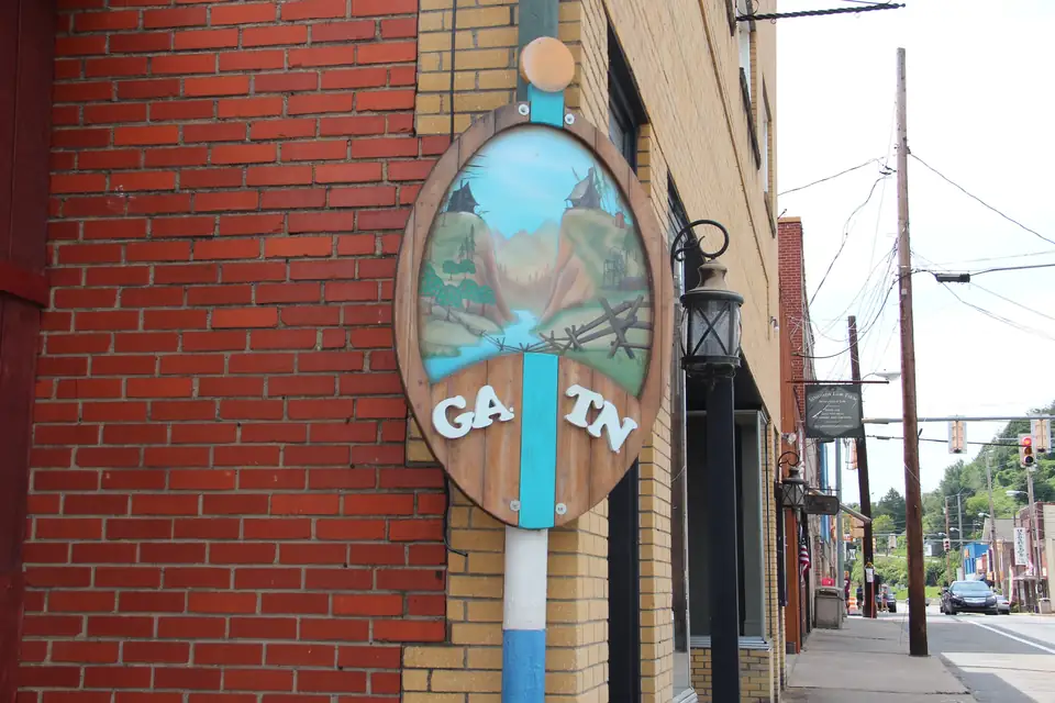 Oval painted wooden sign on a pole against a red-brick storefront, showing a mountain-and-river painting with the letters GA and TN on either side of a blue stripe, with a sidewalk, street lamps and utility poles receding down the street.
