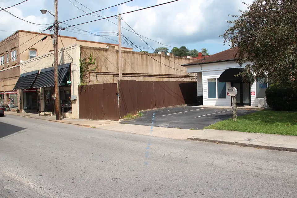 Quiet small-town street with a faint painted blue line crossing the pavement toward a small white building, older brick and stucco storefronts with dark awnings on the left, a wooden fence, utility poles and a partly cloudy sky.