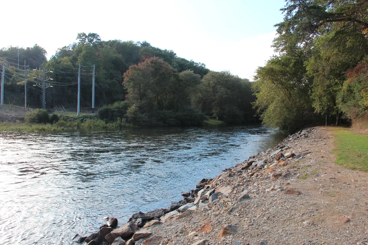 Clear, rippling river water curving along a rock-armored bank with a gravel path at right, green trees beginning to turn color on the far side and a power line with poles crossing the background.