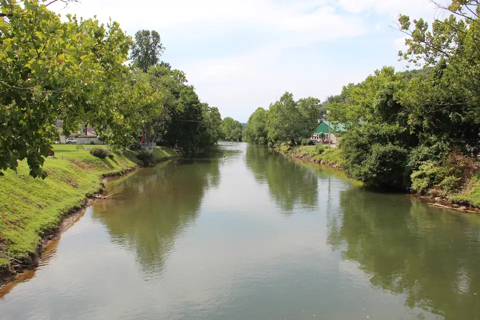 Wide, calm green river reflecting the trees, grassy mowed bank on the left with a house, leafy trees lining both banks, and a green-roofed building on the right bank under a pale blue sky.