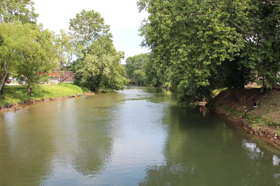 Calm olive-green river between tree-lined banks, a grassy lawn at left with distant buildings and a red-roofed structure, more trees on the right bank and a small bridge in the far distance.