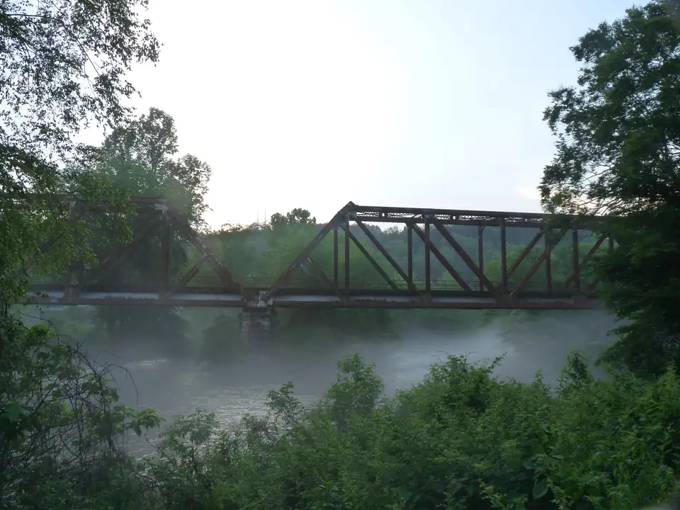 Rusty steel truss railroad bridge spanning the river with low mist rising off the water, framed by dark green trees and leafy brush in the foreground under a bright hazy sky.
